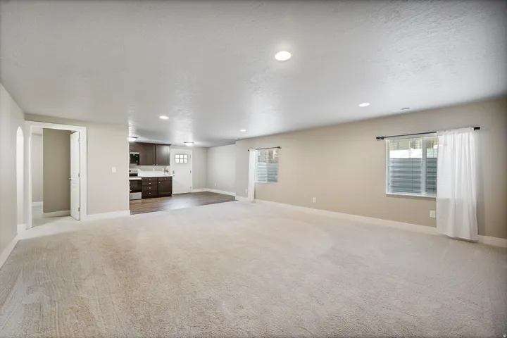 Spacious carpeted area with recessed lighting, two windows, and an adjacent kitchen featuring dark wood cabinetry, white countertops, and stainless steel appliances
