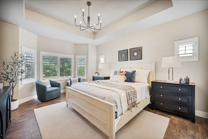 Primary Suite,Tray ceiling with a contemporary chandelier, wood-finish flooring, bay window with plantation shutters, and a paneled accent wall
