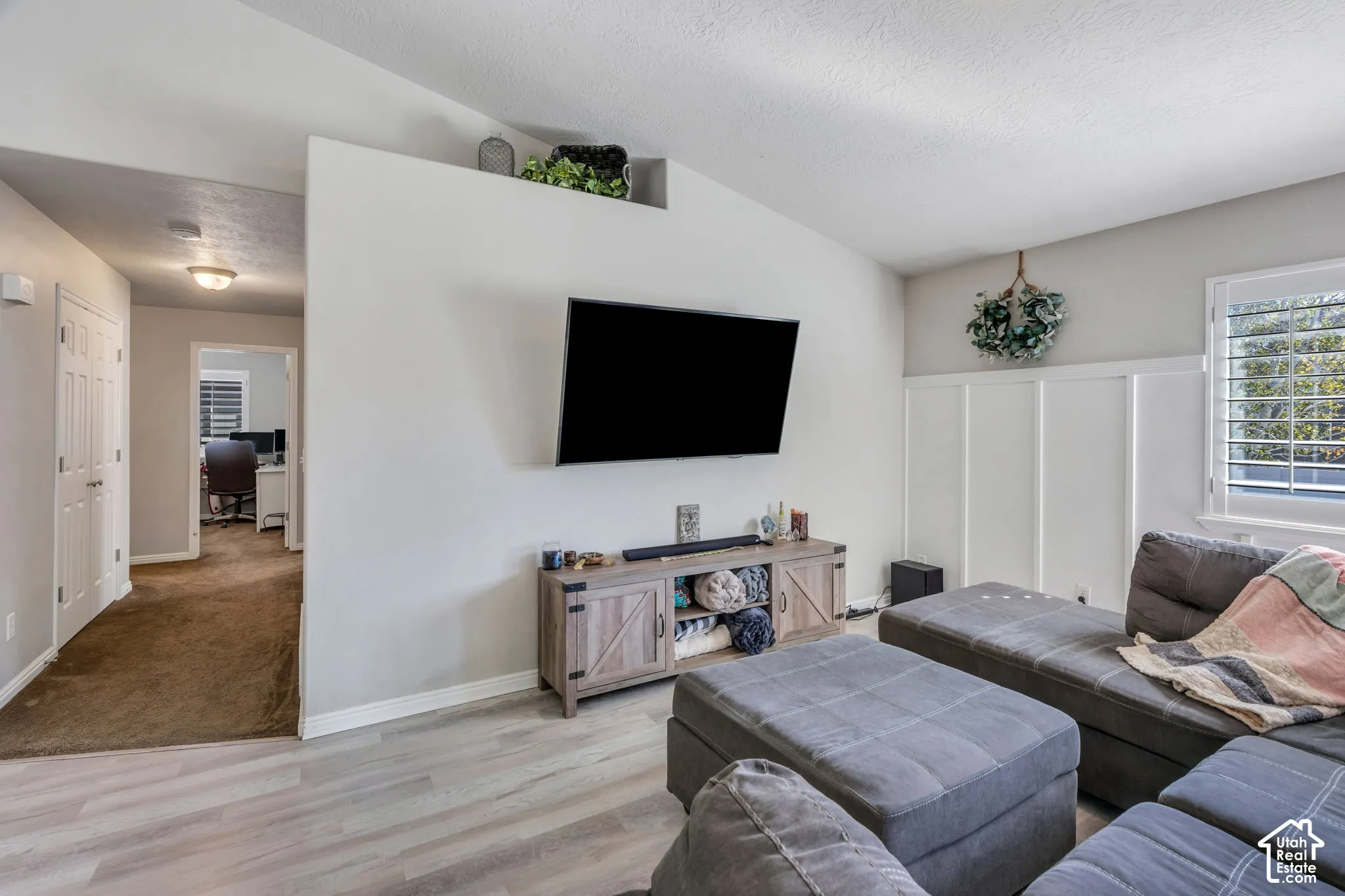 Light wood-finish laminate flooring, accent wall with wainscoting, white window shutters, and a sloped ceiling