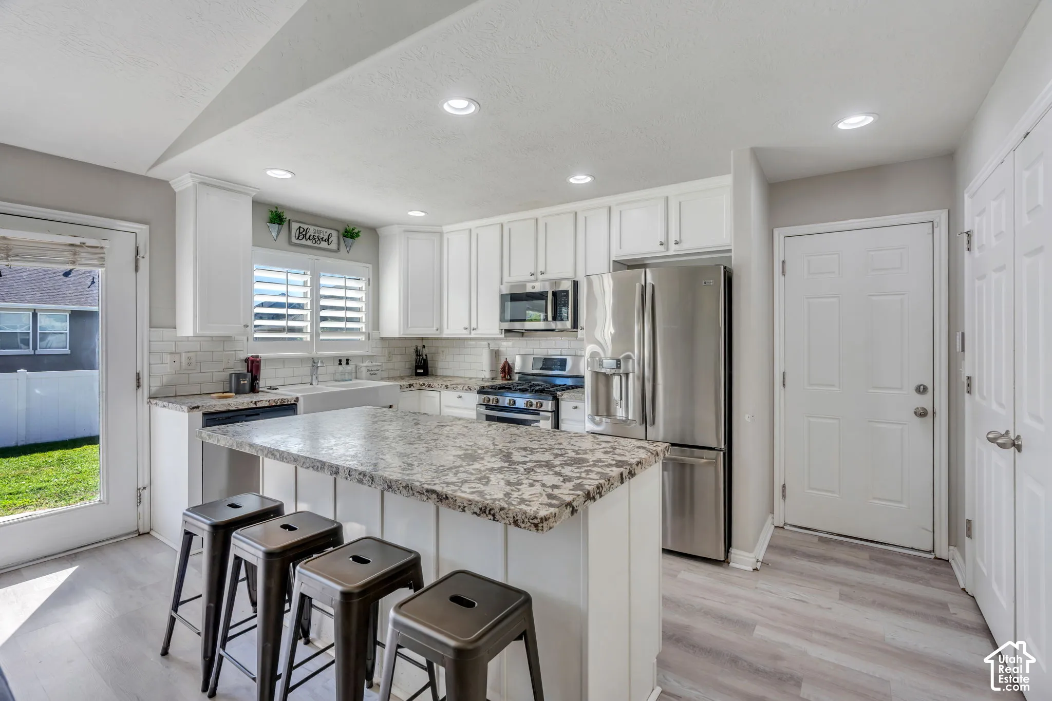 White shaker cabinetry, stainless steel appliances, white subway tile backsplash, light-toned wood-finish laminate flooring, and a large central island