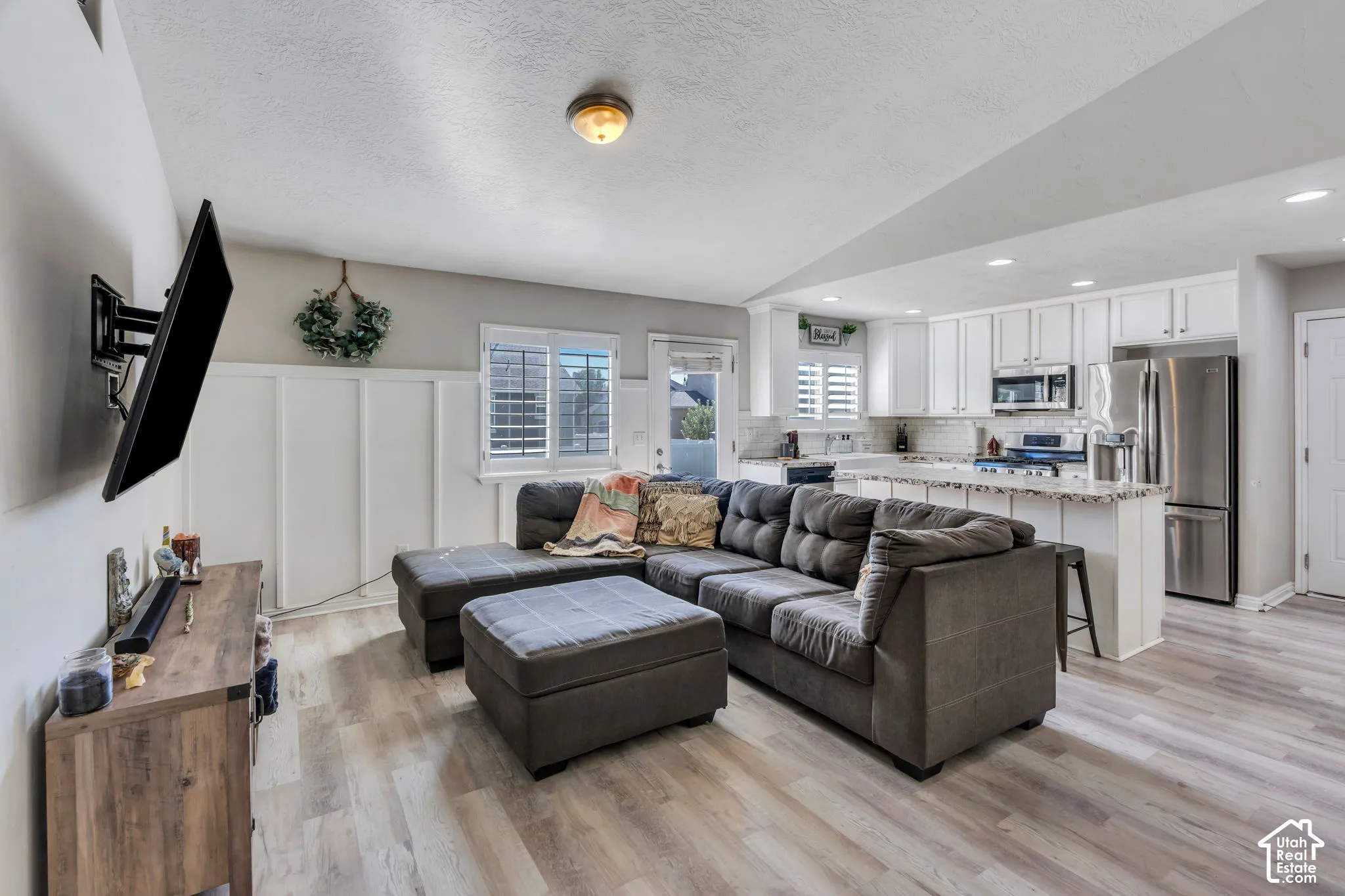 Open-concept living area with wood-finish flooring, board and batten wall detailing, and a kitchen featuring white cabinetry, a tile backsplash, and stainless steel appliances