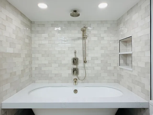Built-in bathtub with a white countertop surround, brushed nickel fixtures, subway tile walls, and a recessed shower niche with shelving