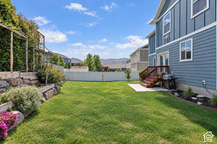 Expansive backyard featuring a manicured lawn, established landscaping, privacy fencing, and a ground-level patio with wooden stairs leading to the home