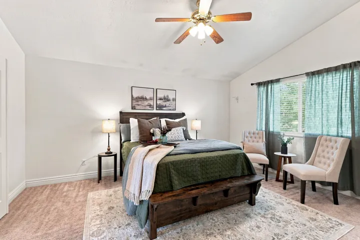 Vaulted ceiling with an attached ceiling fan, large window with blinds, wood-finish headboard, and carpeted flooring