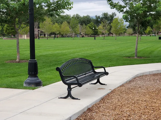 Expansive green space featuring a decorative black metal park bench, paved walking paths, and mature trees