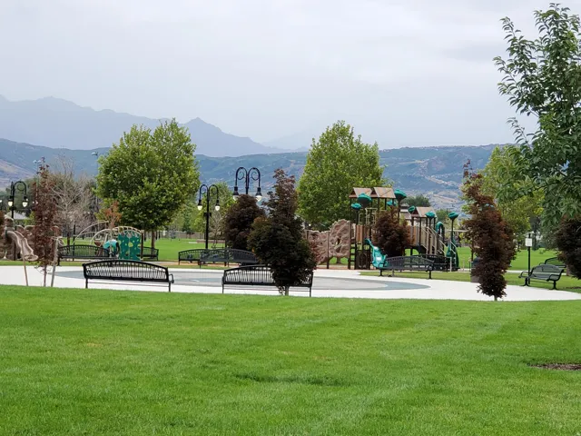 Expansive park area with manicured green space, playground structures, paved walking paths, and mature trees