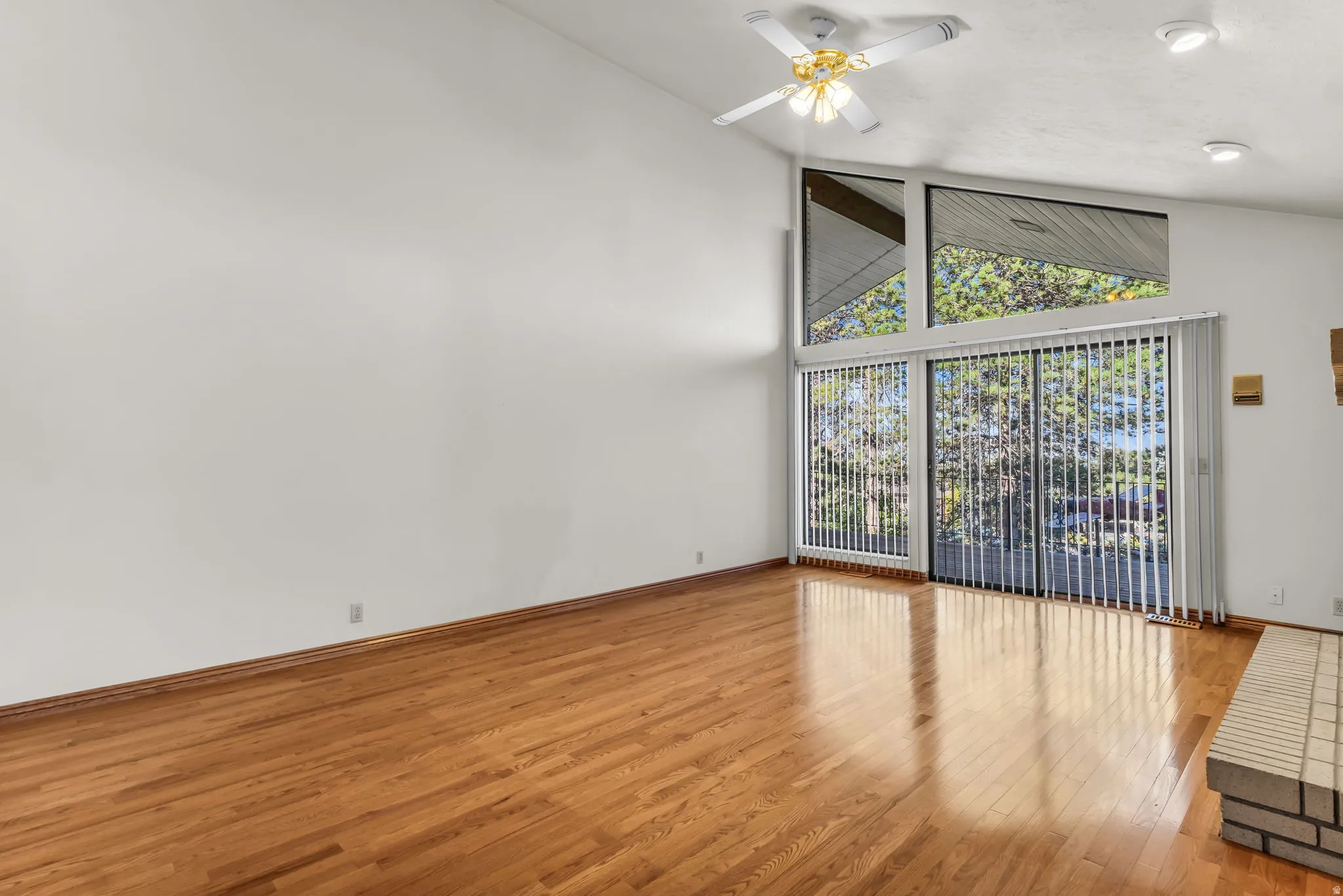 Vaulted ceiling with an integrated ceiling fan, wood-finish flooring, a brick fireplace, and large windows with vertical blinds