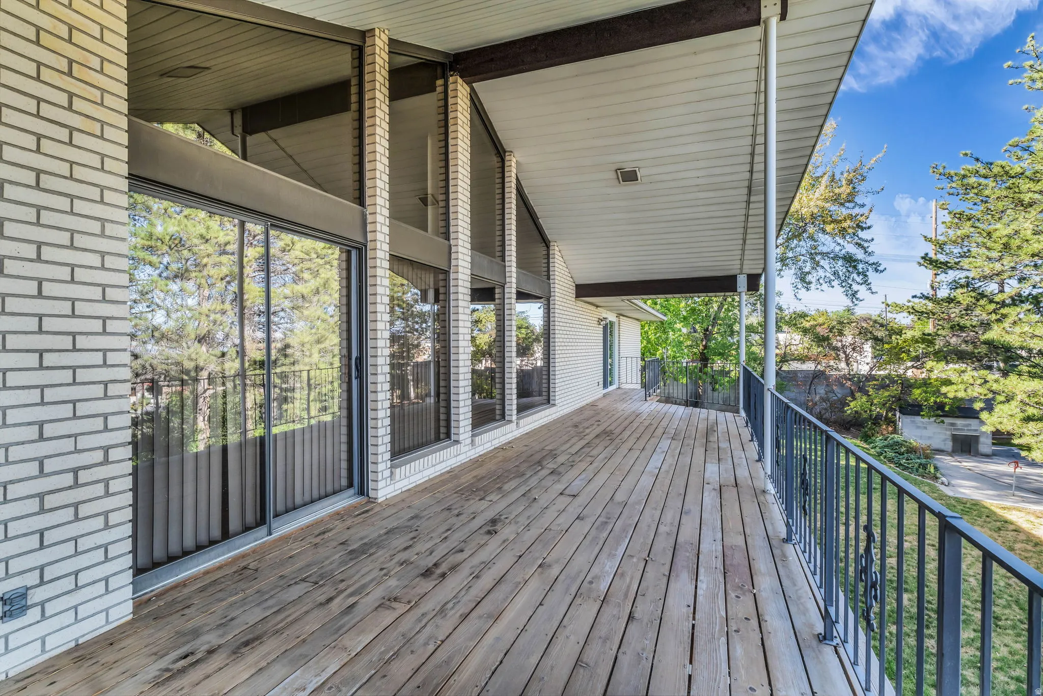 Expansive wood-finish deck with black metal railings, white brick exterior, and full-height windows