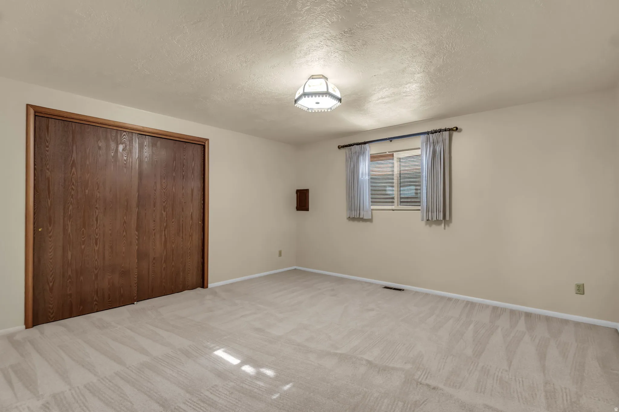 Carpeted flooring, window with blinds, wood-finish sliding closet doors, ceiling-mounted light fixture, and wall-mounted electrical outlet
