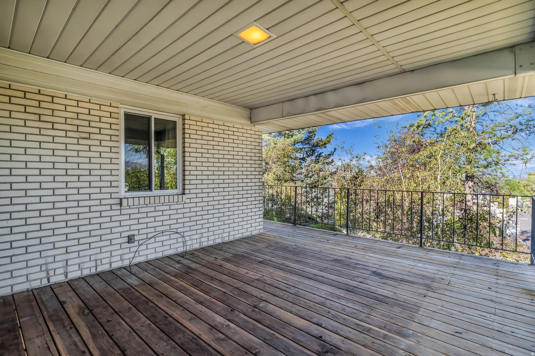 Covered outdoor deck with wood flooring, white brick exterior, black metal railing, and recessed lighting