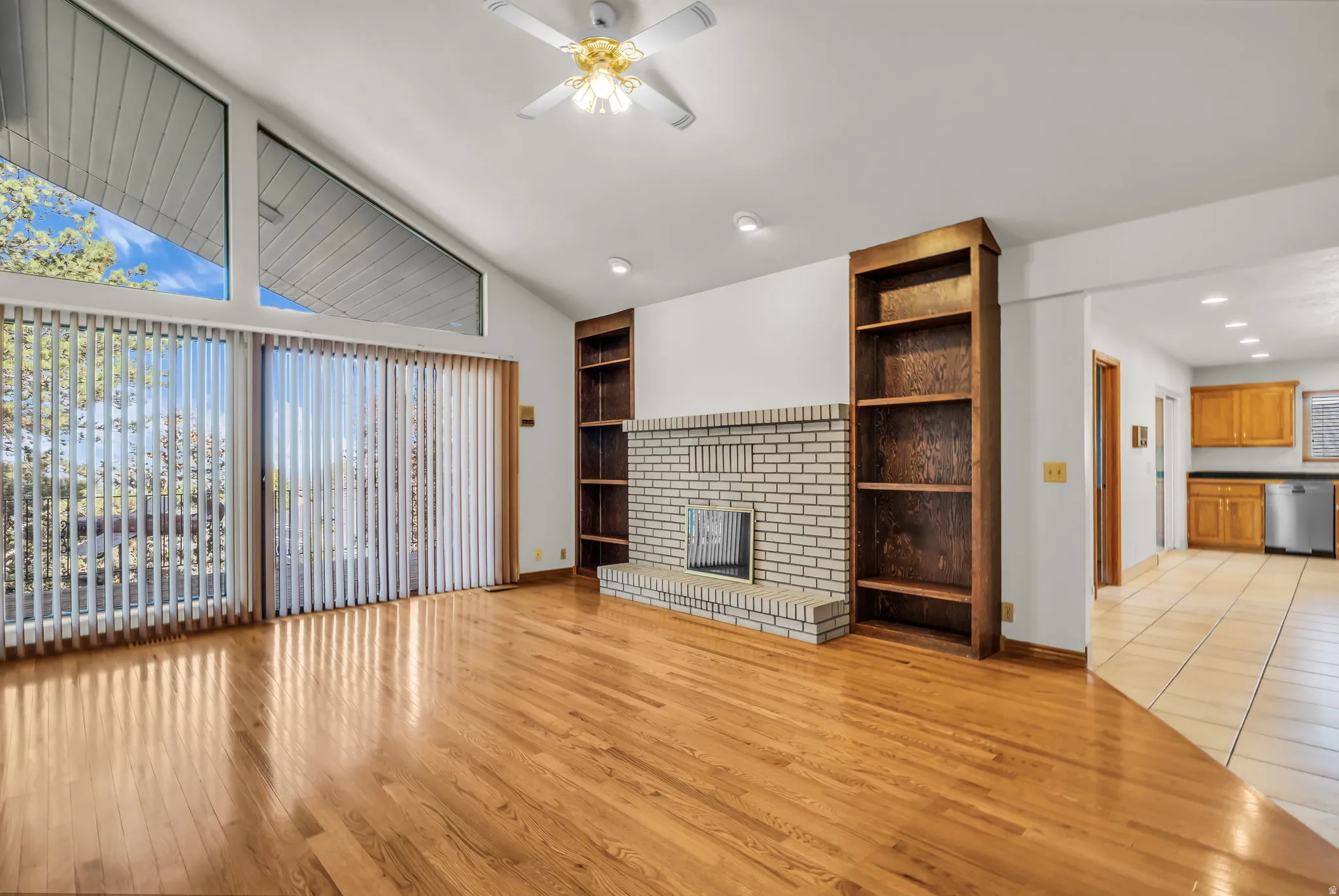 Vaulted ceiling with a ceiling fan, light wood-finish flooring, a white brick fireplace, built-in shelving, and large sliding glass doors