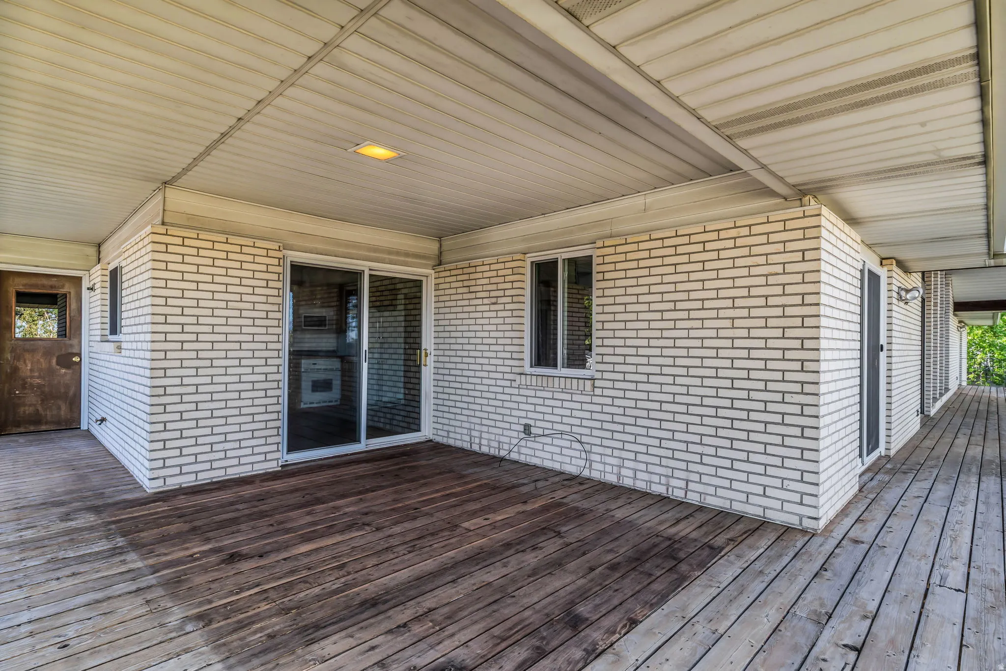 Covered porch featuring wood decking, white brick exterior, a sliding glass door, and a permanent light fixture