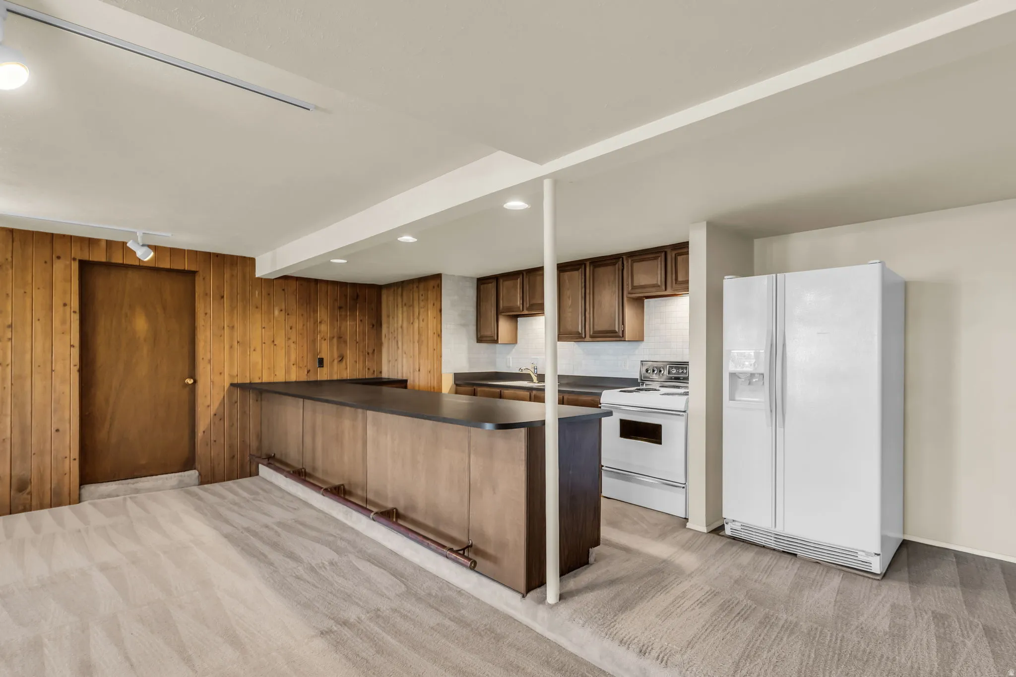 Wood-paneled wall, track lighting, kitchen with dark wood-finish cabinetry, white appliances, and a peninsula with a dark countertop