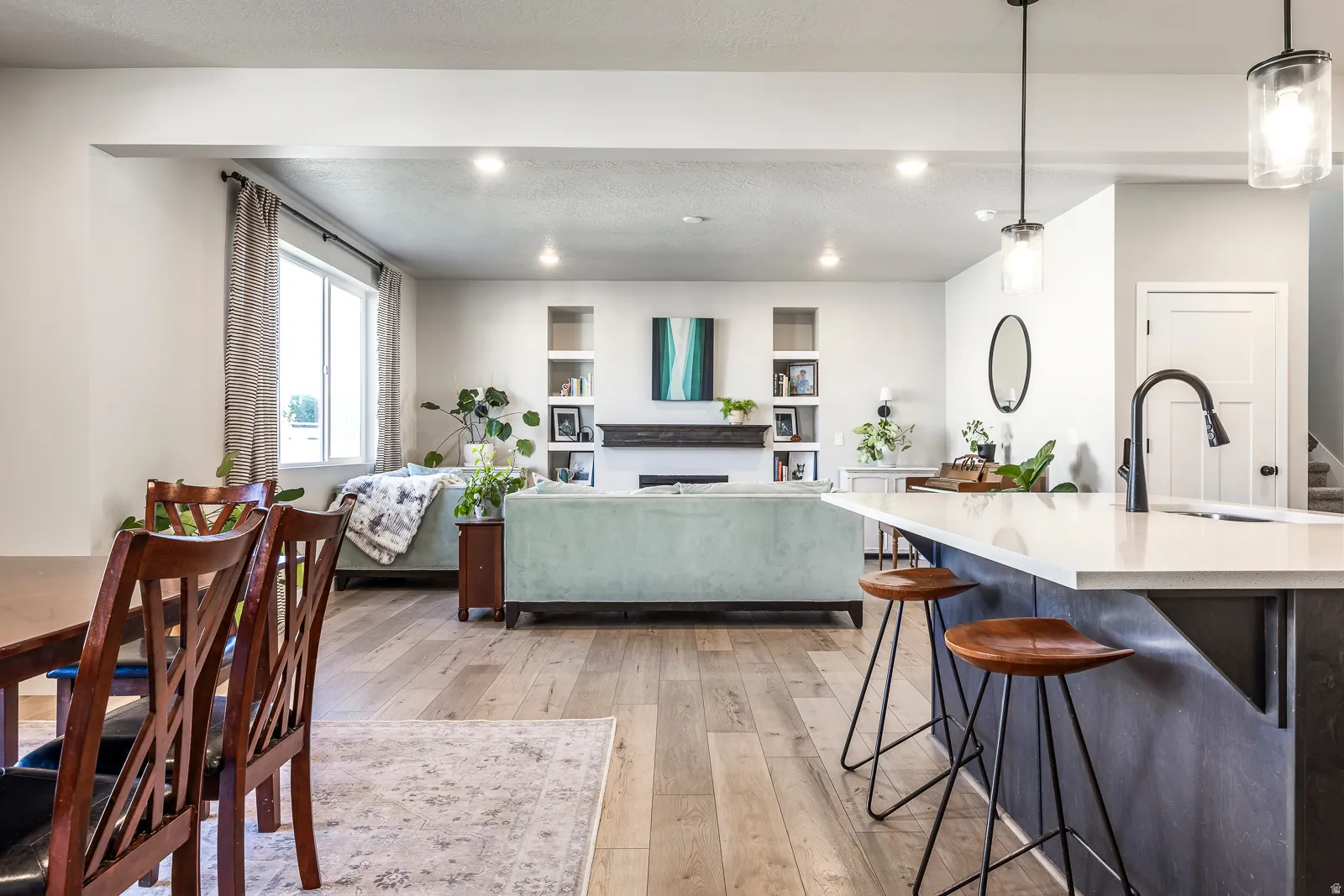 Open-concept living space featuring wood-finish flooring, recessed lighting, a kitchen island with seating, a built-in fireplace with shelving, and large windows