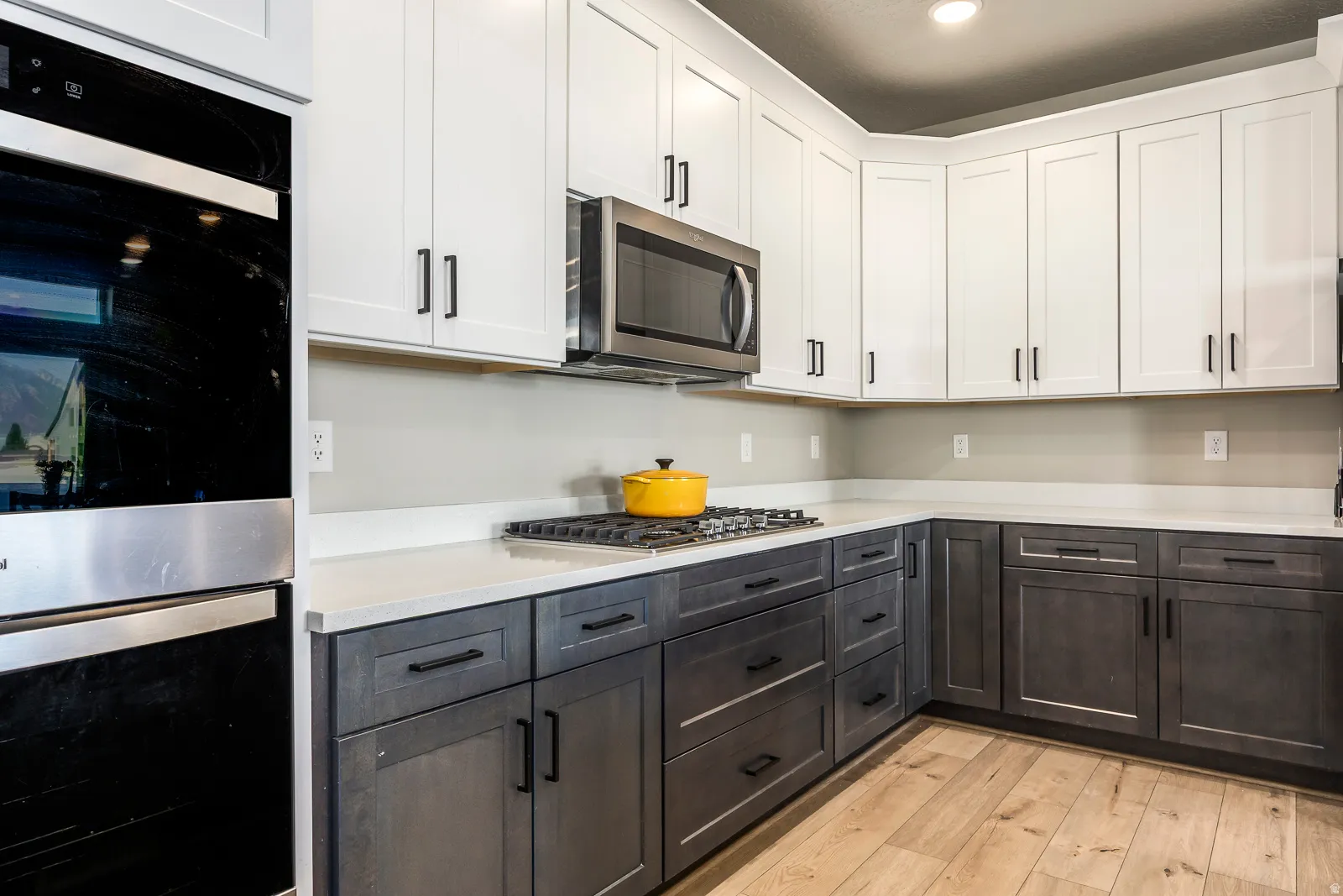 Two-tone cabinetry with white upper cabinets and dark lower cabinets, stainless steel built-in double oven, built-in microwave, gas cooktop, and light-toned wood-finish flooring