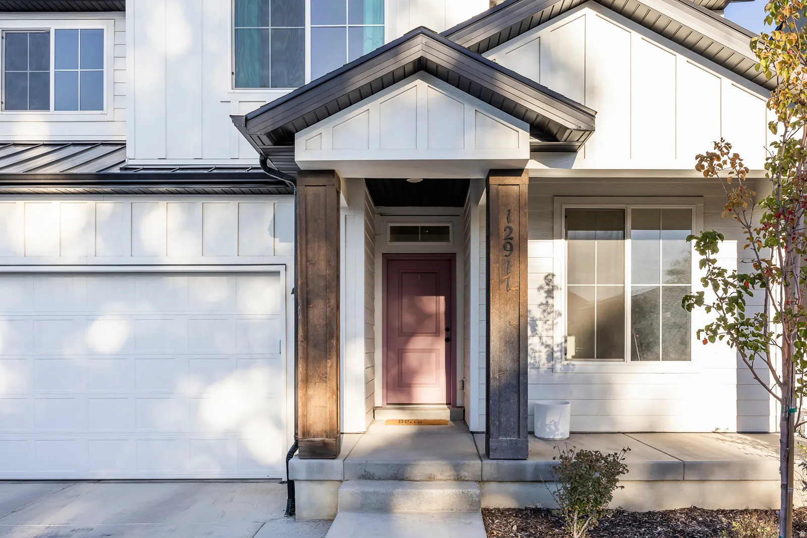 Board and batten siding, dark-framed windows, white panel garage door, covered entryway, and concrete porch