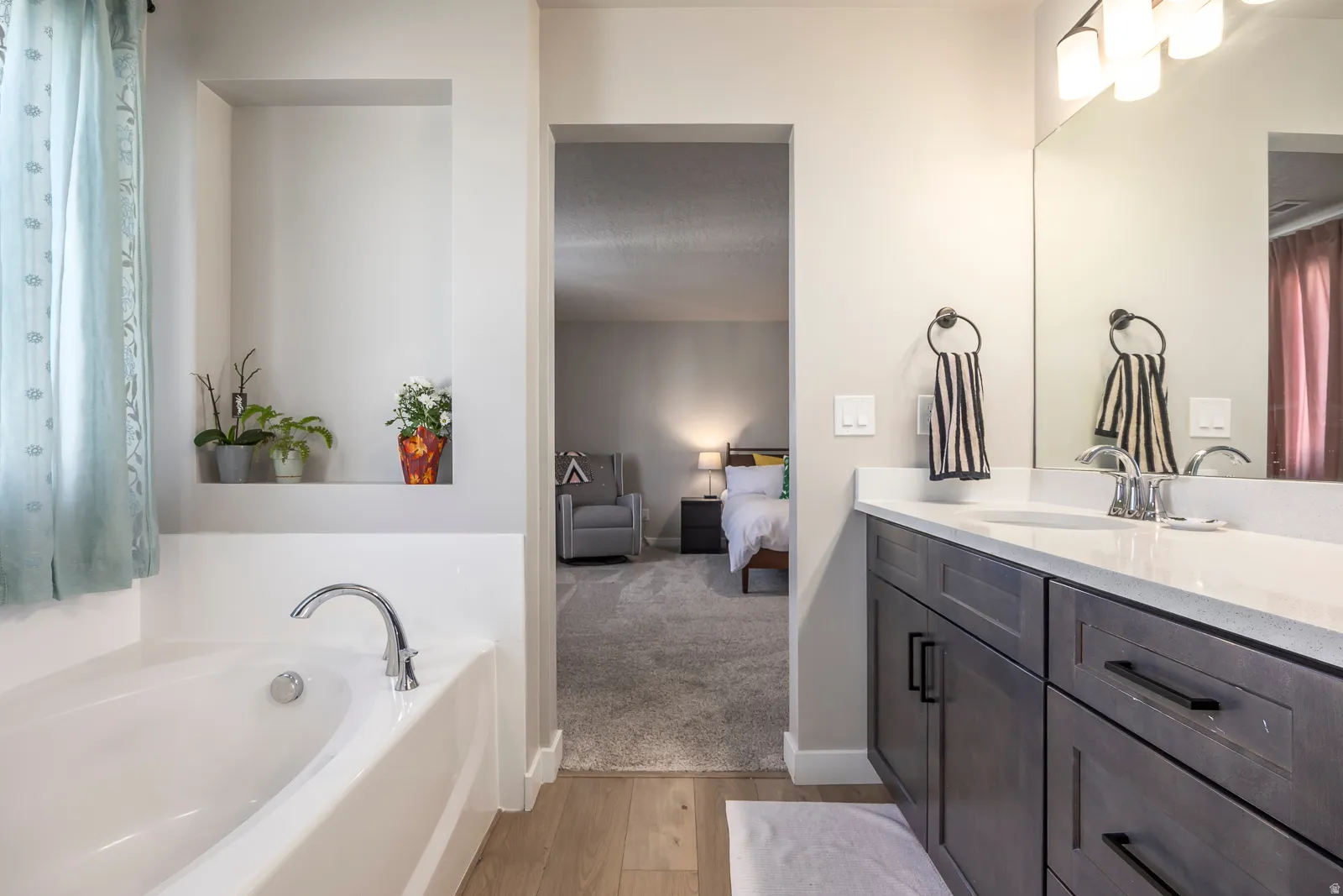 Soaking tub with chrome fixtures, built-in wall niche, double vanity with white countertop, dark wood-finish cabinetry, and large wall-mounted mirror
