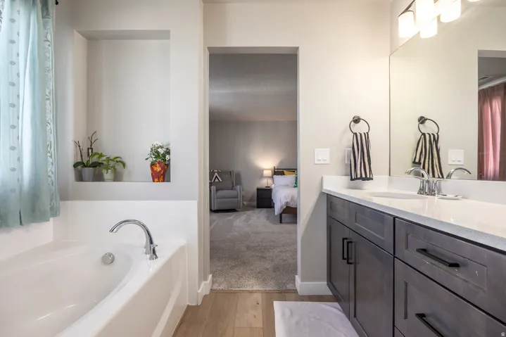Soaking tub with chrome fixtures, built-in wall niche, double vanity with white countertop, dark wood-finish cabinetry, and large wall-mounted mirror
