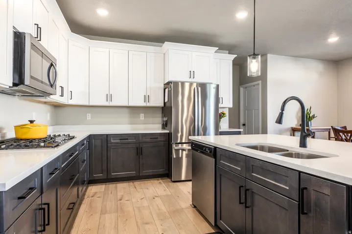 Two-tone cabinetry with contrasting finishes, light-toned countertops, stainless steel appliances, a center island with an integrated sink, and wood-finish flooring