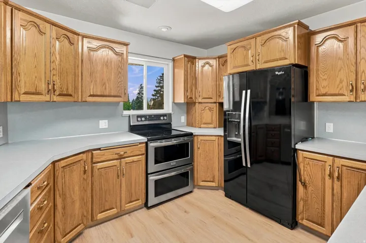 Wood-finish flooring, light wood cabinetry, light grey countertops, stainless steel double oven, and black side-by-side refrigerator