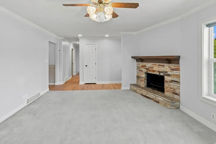 Stone-clad fireplace with a wood mantle, ceiling fan with integrated lighting, wood-finish flooring, and crown molding