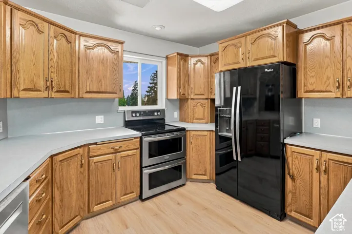 Wood-finish flooring, light wood cabinetry, light grey countertops, stainless steel double oven, and black side-by-side refrigerator