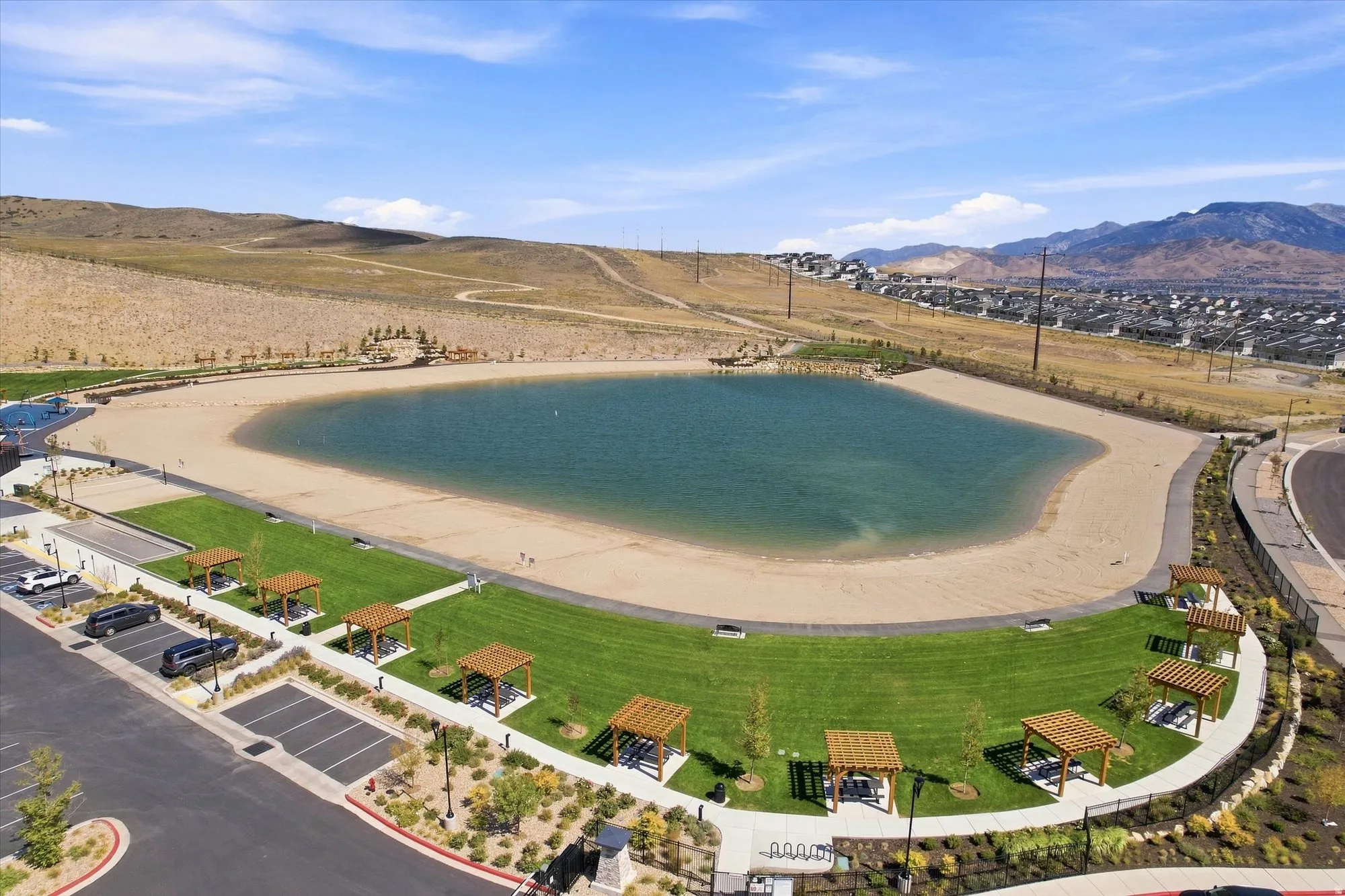 Expansive recreational area featuring a large pond with a sandy beach, a surrounding grassy lawn, multiple pergolas, and a paved parking area