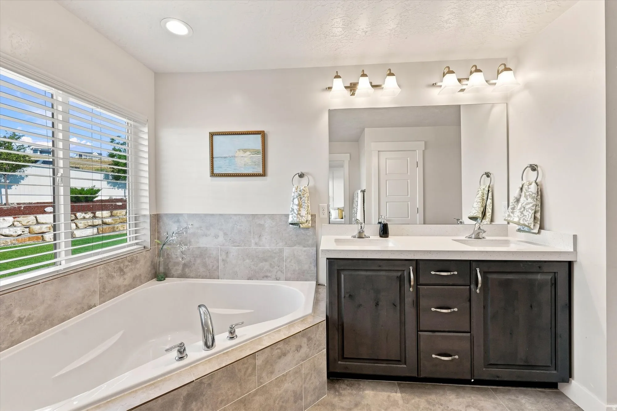 Built-in soaking tub with tile surround, dual sink vanity with white countertop, dark wood-finish cabinetry, and a large wall mirror
