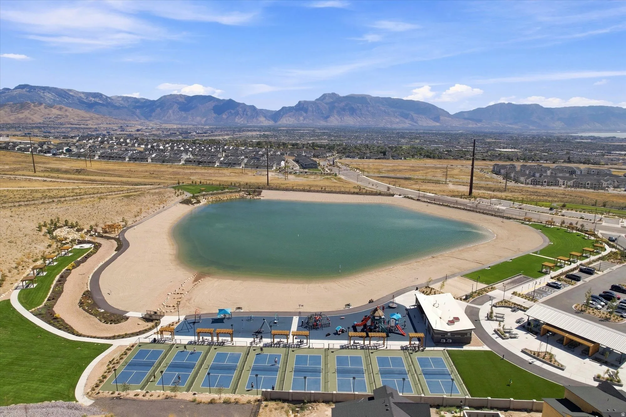 Expansive community lake with a sandy beach, multiple pickleball courts, children's play area, and mountain backdrop