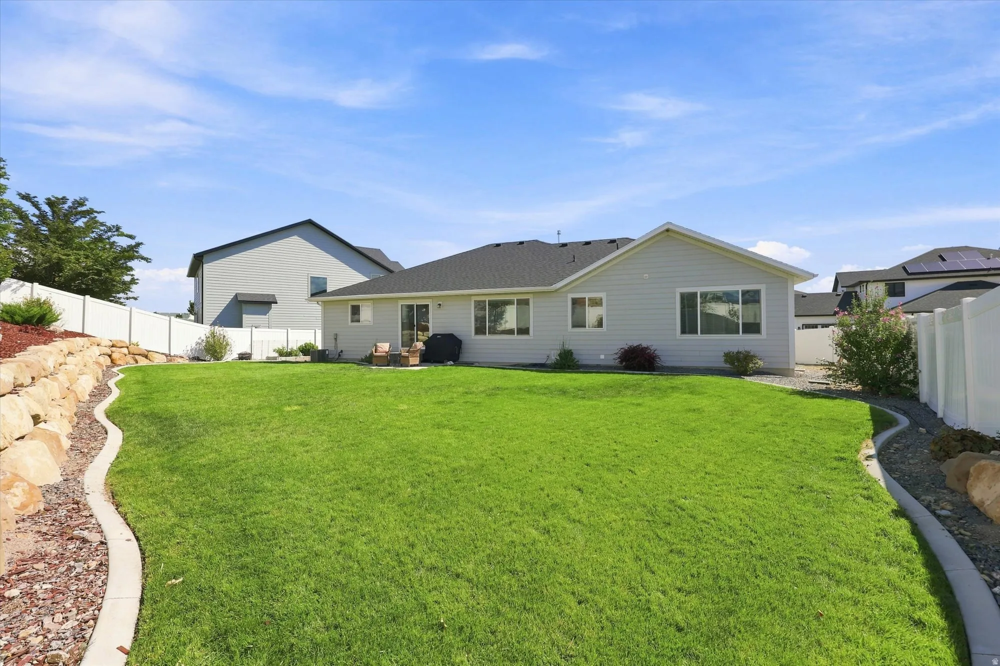 Expansive backyard featuring a manicured lawn, concrete curbing, a rock wall with landscaping, and white privacy fencing