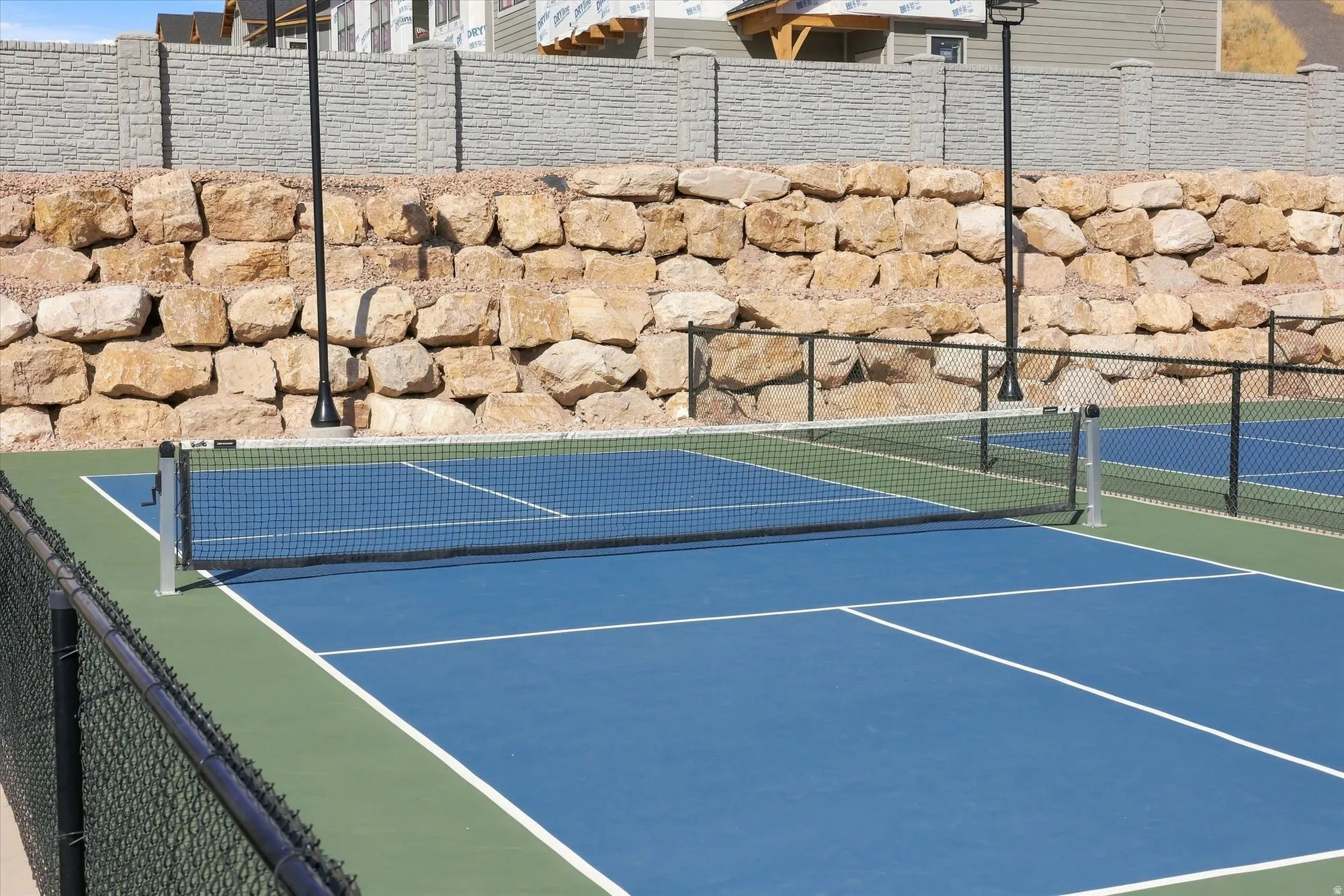 Outdoor court featuring blue and green playing surfaces, white court lines, a black net, and perimeter fencing