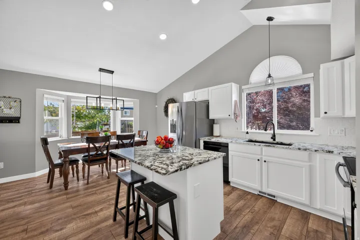 Vaulted ceilings with recessed lighting, wood-finish flooring, white cabinetry, a kitchen island with stone countertop, and a bay window