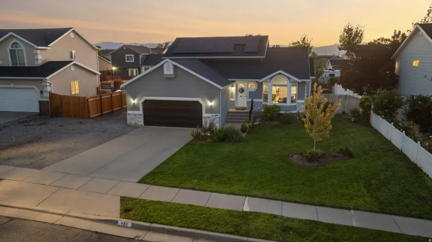 Gray exterior with stone accents, two-car garage, manicured lawn, solar panels, and white picket fencing