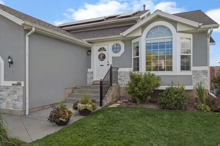 Stucco exterior with stone accents, bay window with arched transom, solar panels, white entry door with an octagonal window, and concrete entry steps with a black railing