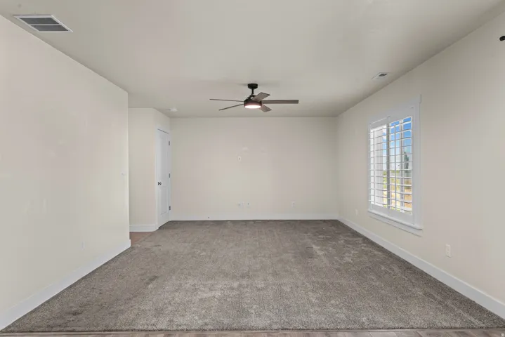 Expansive room featuring neutral-toned carpeting, wood-finish flooring, a ceiling fan, and a window with white louvered shutters