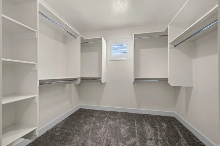 Built-in white shelving, multiple clothing rods, window with horizontal blinds, gray carpeting, and white wall finishes