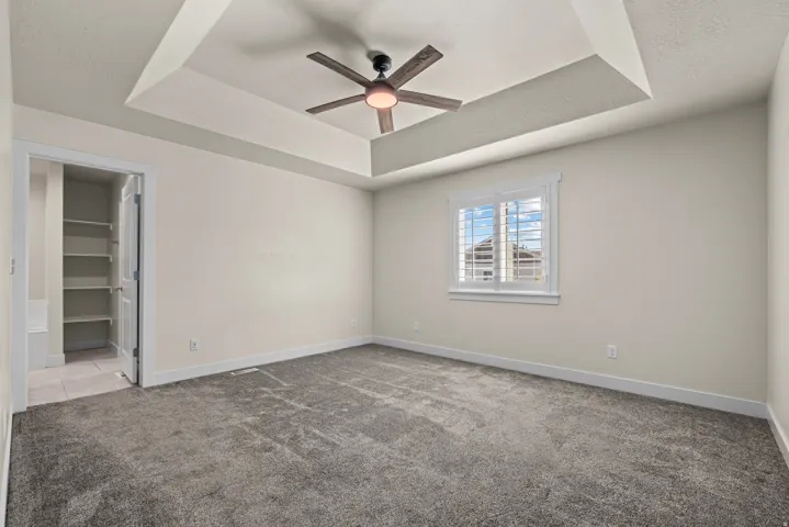 Tray ceiling with a ceiling fan, window with plantation shutters, gray carpet, and an open doorway revealing built-in shelving