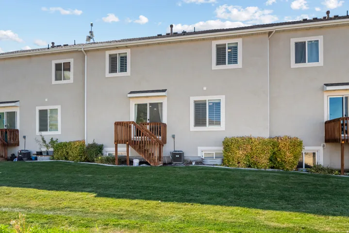 Stucco exterior with multiple windows, an upper-level deck, and a ground-level patio, set on a manicured lawn