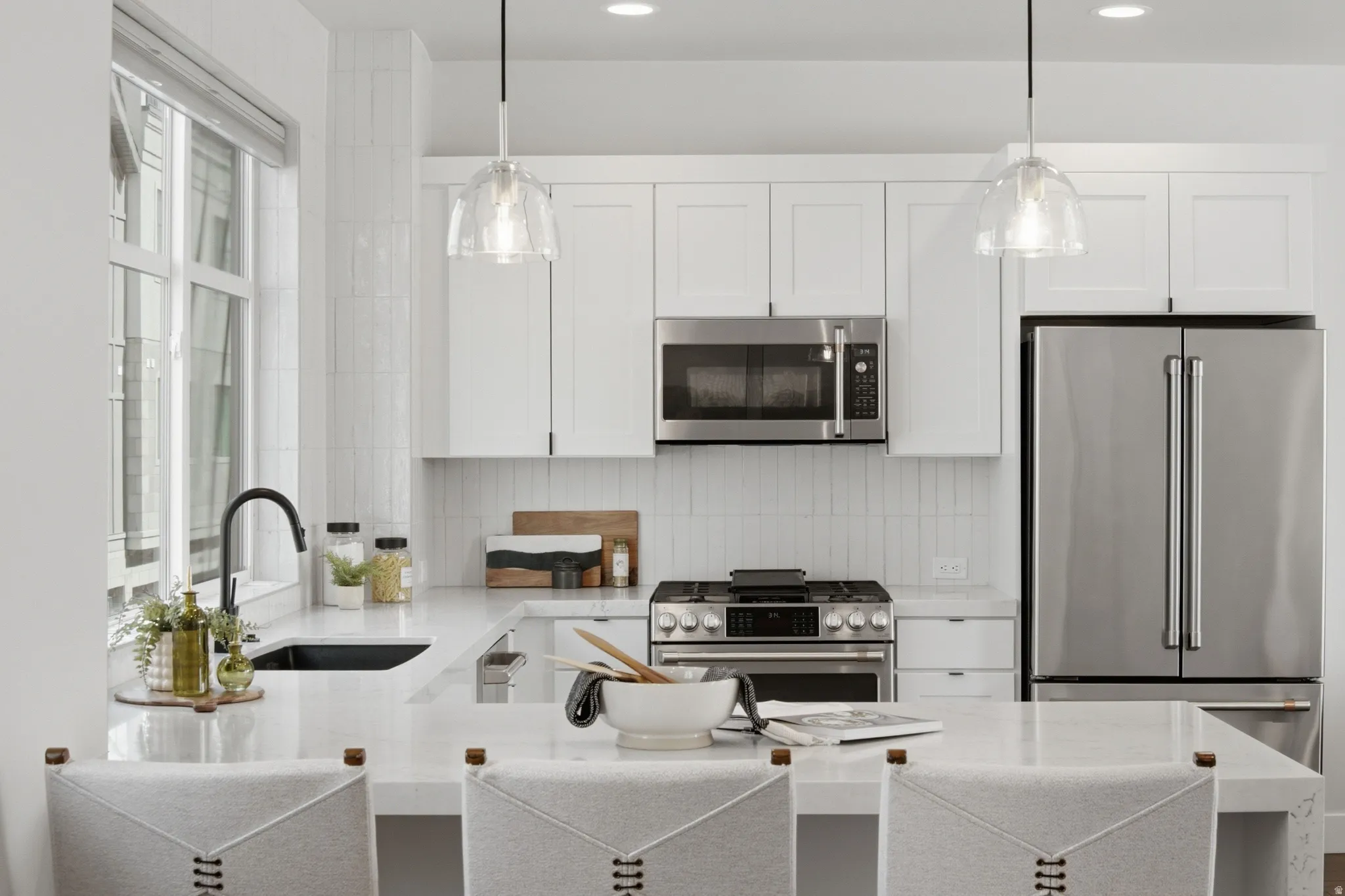Well-appointed kitchen featuring white cabinetry, stainless steel appliances, a large window, a black sink, and pendant lighting