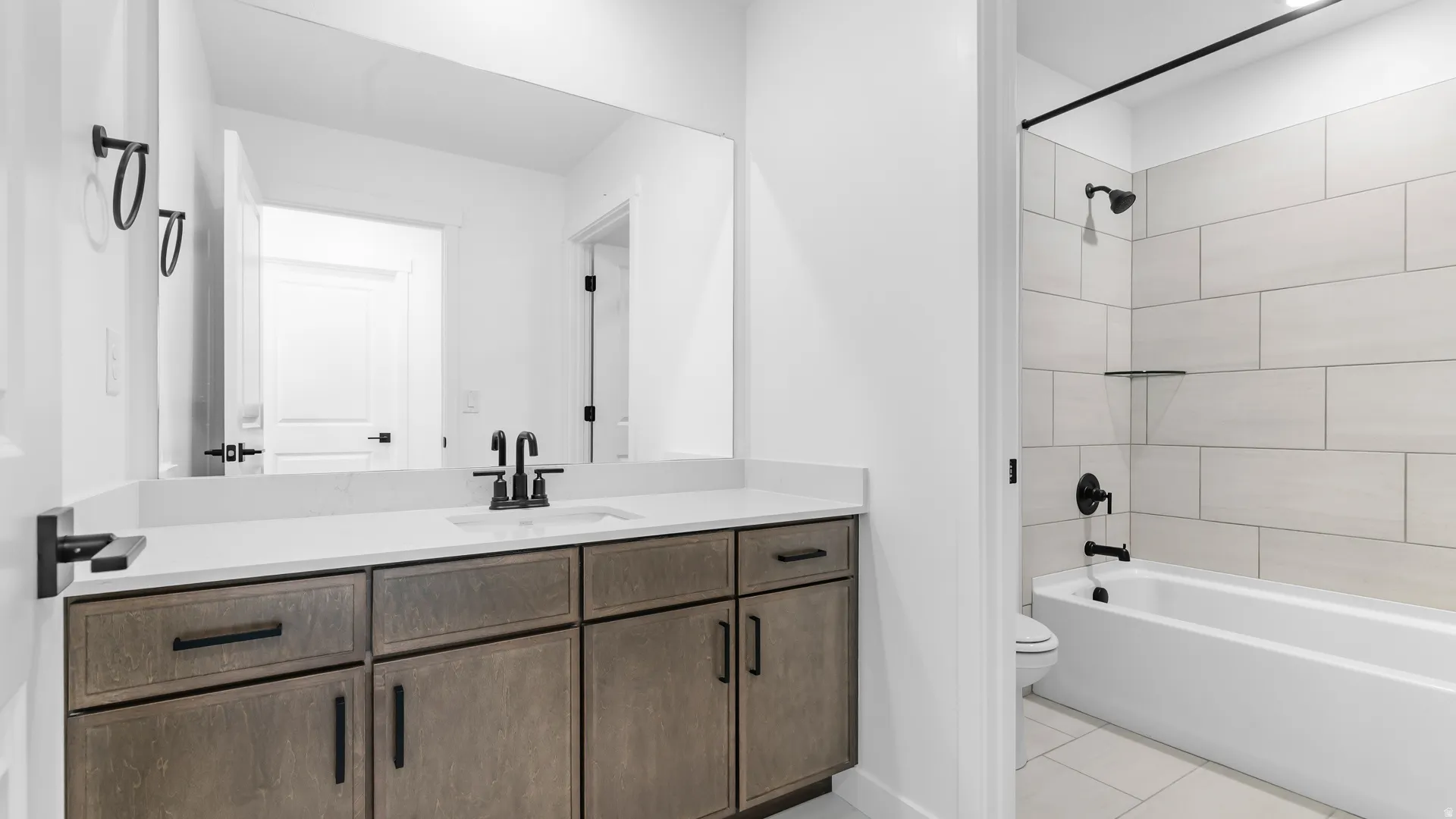 Bathroom featuring a wood-finish vanity with a light-toned countertop, a black-finished faucet, and a shower-over-tub combination with large format wall tiles