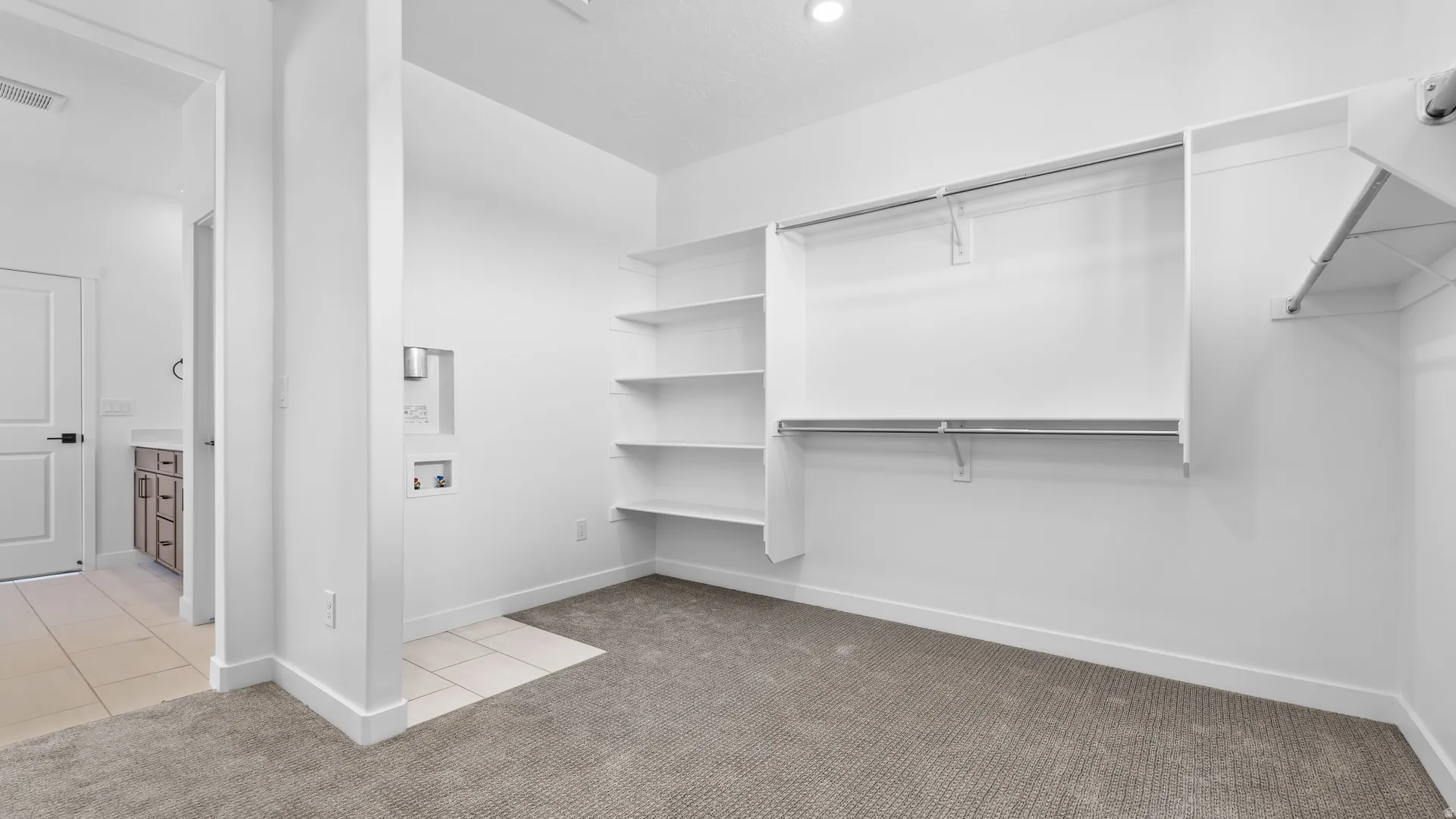 Wall-mounted shelving, built-in clothing rods, and soft carpeting complement the laundry connections in this versatile space