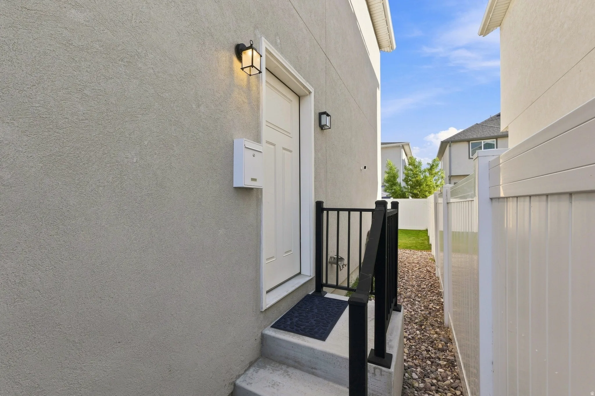 Stucco exterior, white paneled entry door, black metal railing, gravel pathway, and white privacy fence