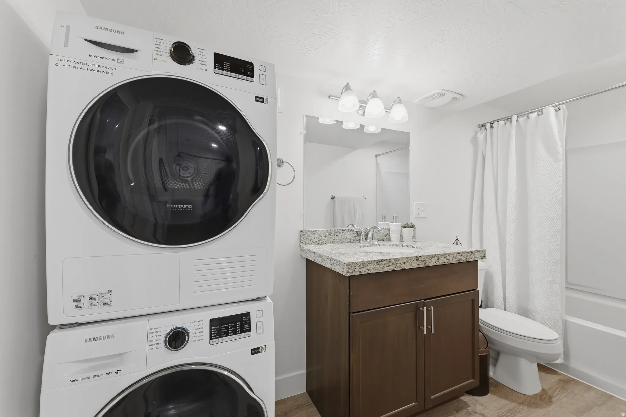 Stacked washer and dryer, vanity with a stone-finish countertop, undermount sink, wall-mounted light fixture, and a tub with shower