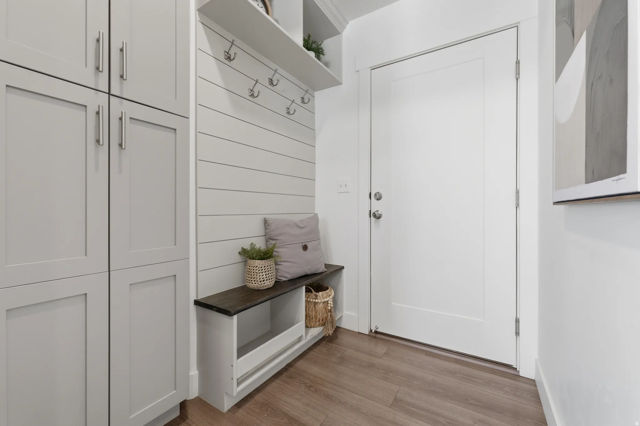 Built-in cabinetry with ample storage, wood-finish flooring, white shiplap wall accent, integrated bench with a dark wood top, and wall-mounted coat hooks