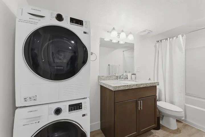 Stacked washer and dryer, vanity with a stone-finish countertop, undermount sink, wall-mounted light fixture, and a tub with shower