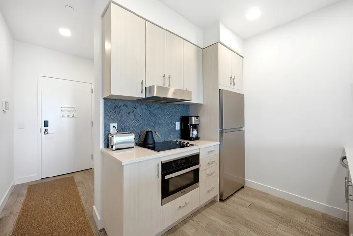 Kitchenette featuring light wood-finish cabinetry, stainless steel appliances, a built-in cooktop, and a blue herringbone tile backsplash