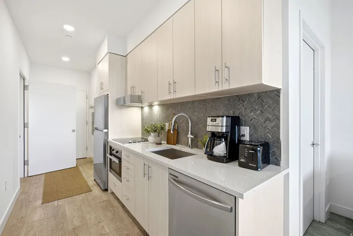 Galley kitchen with wood-finish cabinetry, stainless steel appliances, light-toned countertop, and herringbone tile backsplash