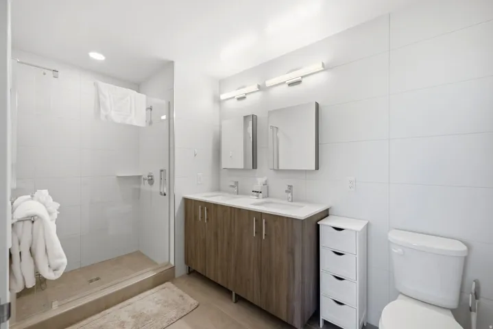 Tiled shower enclosure with a glass door, wood-finish double vanity with dual sinks, and white tile walls