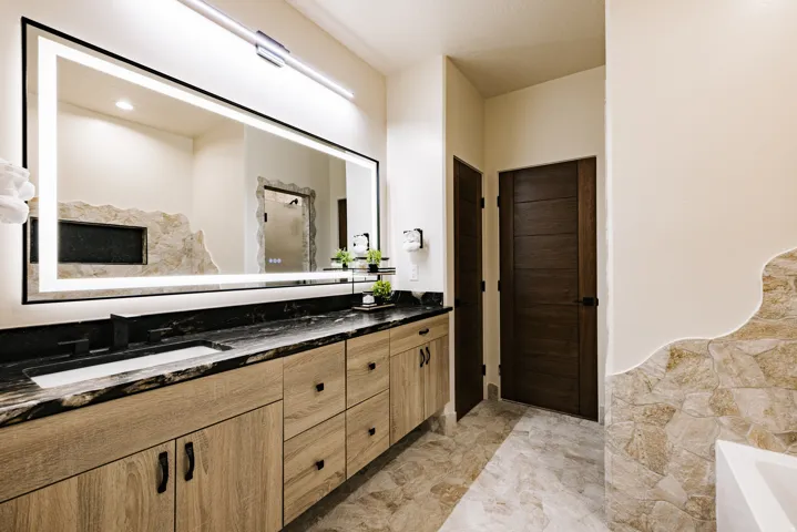 Dual vanity with integrated sinks, black countertop, light wood-finish cabinetry, and a large illuminated mirror