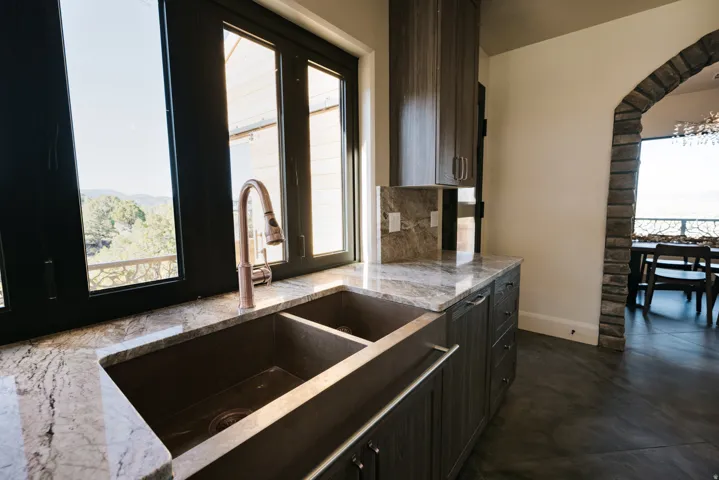 Double basin sink with a rose gold faucet, stone countertops, dark wood-finish cabinetry, and a stone archway